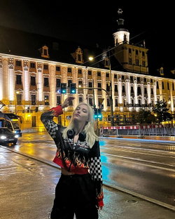 Slim blonde in checkered flame jacket and baggy pants poses on wet urban street at night.