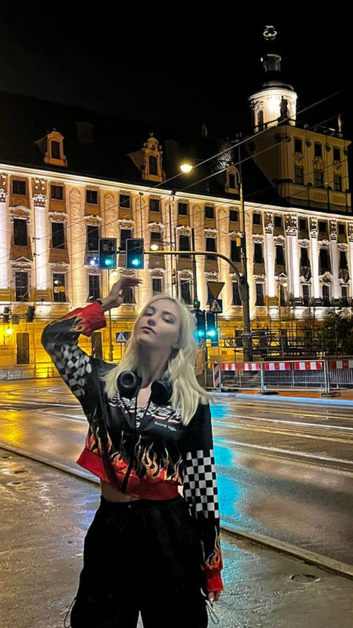Slim blonde in flame checkered racing jacket and black pants poses on wet night street by ornate building.