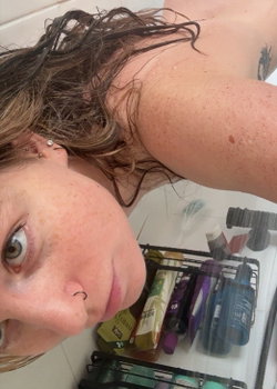 A person with light skin and dark hair lies on a bed with makeup products in the foreground.
