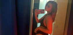 Woman with long brown hair in a black dress taking a selfie in a dimly lit room.