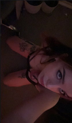A woman with dark hair and visible arm tattoos lies on her side in a dimly lit room, looking directly at the camera in