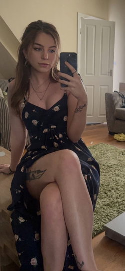 A woman with long brown hair sits on a wooden table in a floral dress, holding her phone for a mirror selfie; visible
