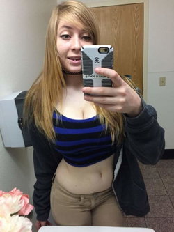 Blonde woman in a striped crop top and beige pants takes a mirror selfie, wearing a dark jacket and choker in a room