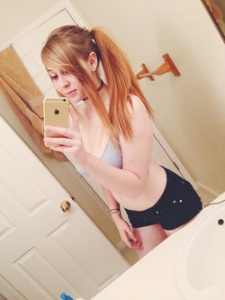 Redheaded woman in a gray bra and black shorts takes a mirror selfie in a bathroom, holding a gold phone.