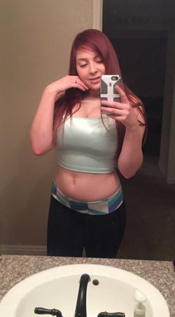 Redhead woman in a light top and patterned skirt taking a selfie in a bathroom.