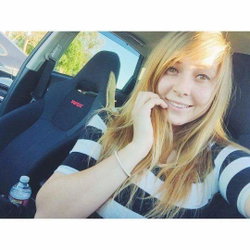 A young woman with long blonde hair sits in a car, smiling and touching her face.