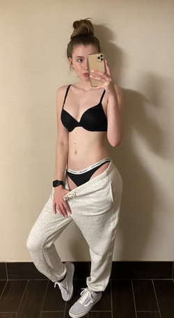 A woman with brown hair in a bun wears a black bra and thong with gray sweatpants pulled down, posing in a mirror