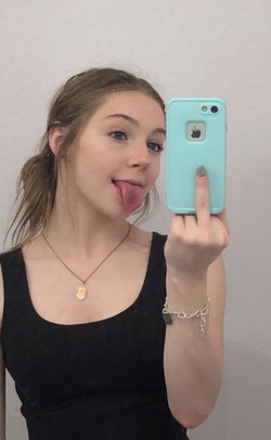 A young woman with light brown hair takes a playful mirror selfie, sticking out her tongue and holding up a blue phone