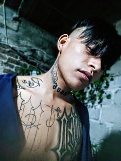 A man with dark hair and multiple tattoos on his neck and chest, wearing a blue open shirt, leaning against a brick