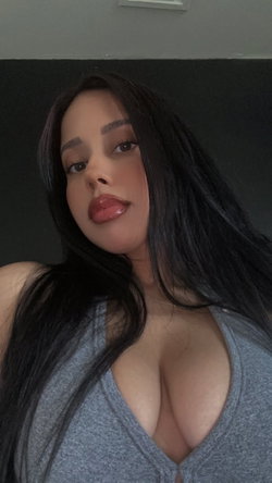 A woman with long dark hair and a curvy build wears a low-cut gray top, posing indoors against a dark wall.