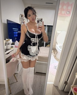 A woman in a black and white lace maid outfit with thigh-high stockings takes a mirror selfie in a bedroom with visible