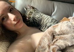 A woman with long brown hair lies topless on a couch, smiling softly at the camera, with a tabby cat resting on her