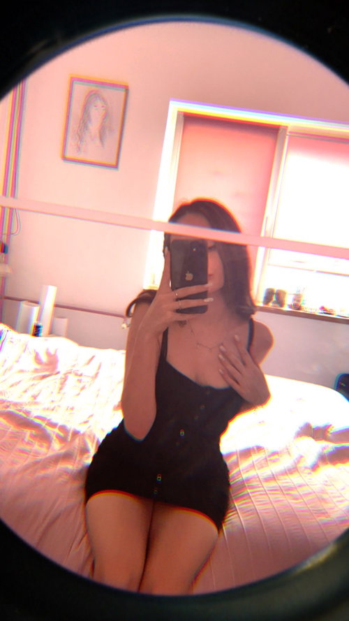 A woman with dark hair takes a mirror selfie in a black button-down dress, sitting on a bed in a brightly lit room with