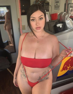 Curvy woman in red bikini poses indoors, showing rose tattoos on hips and holding a red strap.