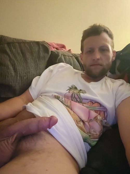 A man with short hair and a beard lies on a couch, wearing a white graphic t-shirt and dark pants, with his hand on his