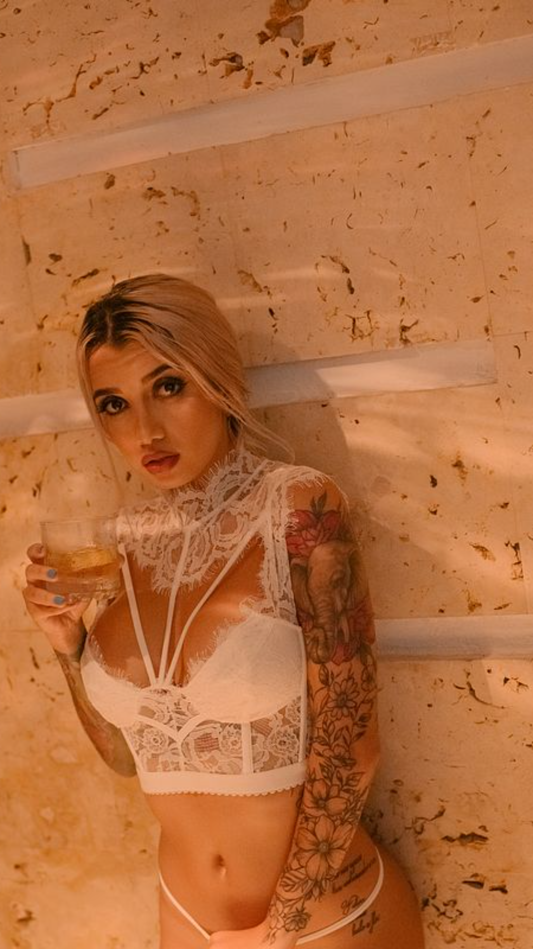 Tattooed blonde in white lace lingerie holding a drink against a stone wall.