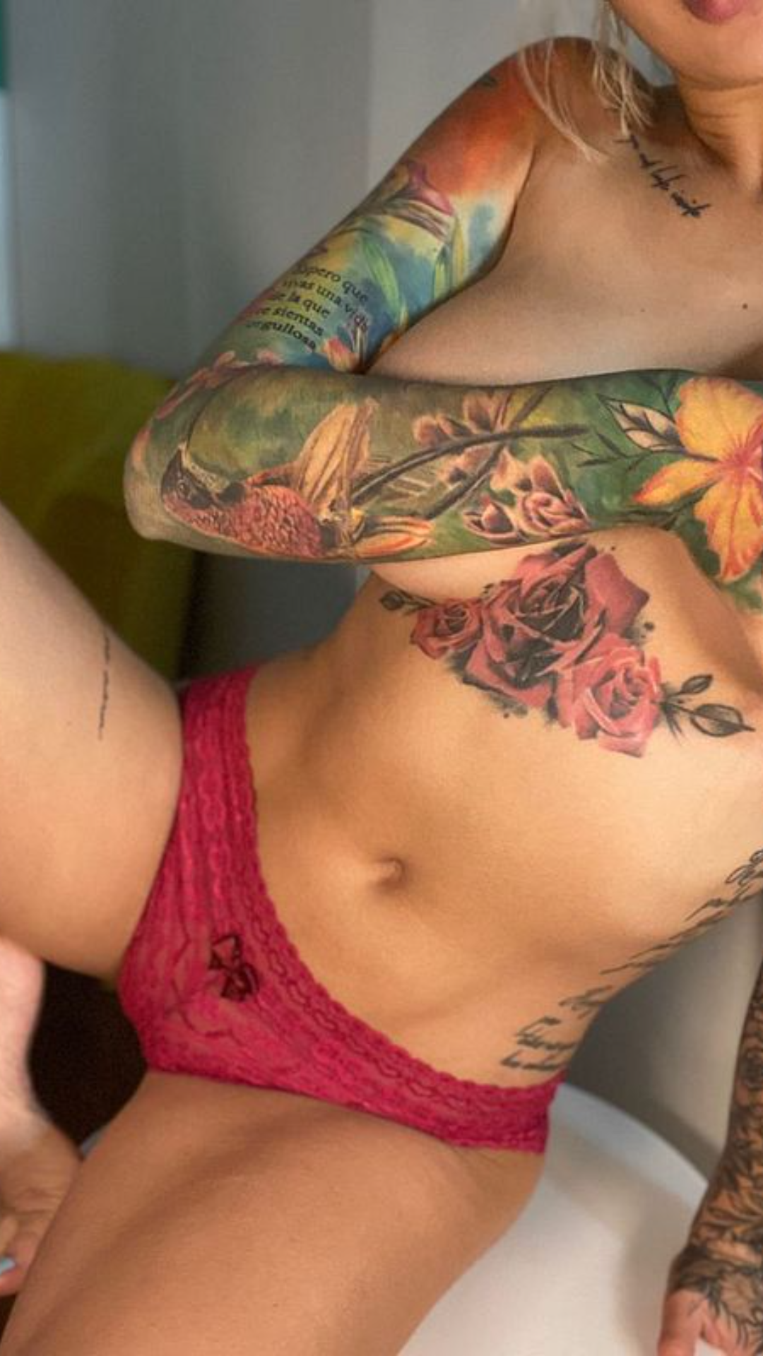Blonde woman with extensive colorful tattoos wearing only pink lace underwear in an indoor setting.