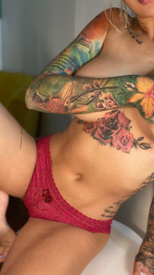 Blonde woman with extensive colorful tattoos wearing only pink lace underwear in an indoor setting.