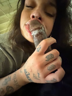 Person with dark hair and tattoos on arms licking a large lollipop.