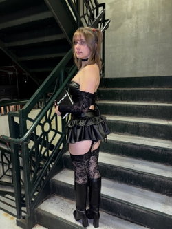 A woman with blonde hair in a ponytail stands on outdoor stairs wearing a black leather corset, lace sleeves, a short