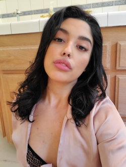 Brunette woman with wavy hair and nose ring, wearing a light pink robe over black mesh lingerie, posing in a bathroom