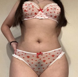 Woman with curvy figure wearing sheer red heart-patterned lingerie with white lace trim, posing indoors.