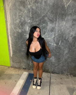 A curvy woman with long black hair poses against a gray wall, wearing a black top and denim skirt, making a playful