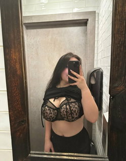 Woman with long dark hair in a black lace bra and black pants taking a selfie in a bathroom mirror.