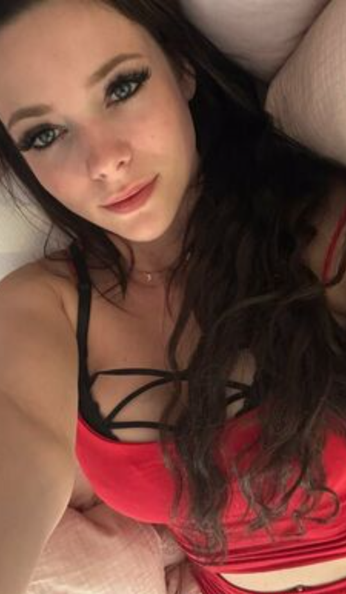 Brunette woman with long dark hair lying down, wearing a red and black strappy top, looking at the camera in a close-up