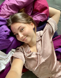 A young woman with brown hair lies on colorful blankets, wearing a silky pink shirt, smiling softly at the camera.