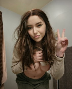 A woman with long brown hair and a nose piercing poses in a room, pulling down her sweater to expose her bare chest