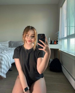 A young woman with blonde hair and a black dress sits on a bed taking a selfie.