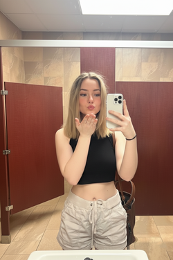 Blonde woman in black crop top and light shorts taking a selfie in a bathroom.