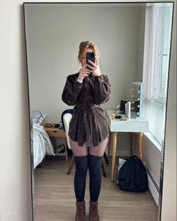 Blonde woman in a brown coat and pink shorts taking a selfie in a bedroom.