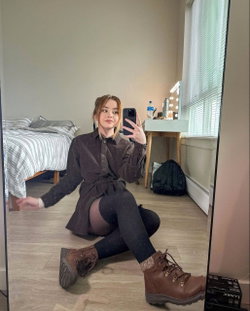 A young woman with blonde hair sits on the floor in a cozy room, taking a selfie in the mirror.