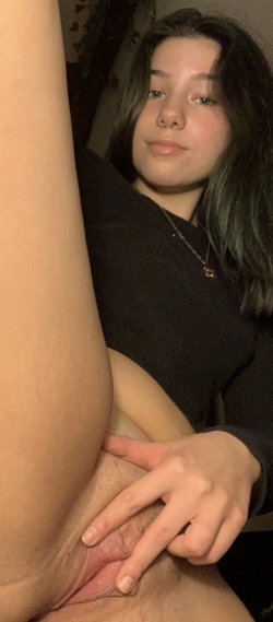 A woman with dark hair and a nose ring in a black top is holding a man's erect penis in a dimly lit indoor setting.