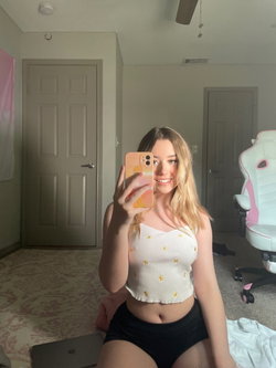 Blonde woman in a white ribbed crop top and black shorts taking a selfie in a bedroom.