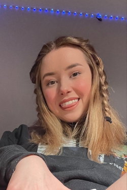 A smiling woman with blonde braided hair, wearing a dark hoodie, sitting indoors with blue string lights above her.
