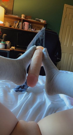 Person in gray socks and black pants sits on a bed in a cluttered room.