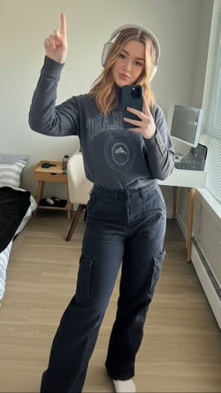 Blonde woman in gray sweatshirt and dark cargo pants takes a mirror selfie with headphones on, pointing upward in a