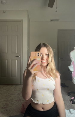 Blonde woman in a white floral crop top and black shorts takes a mirror selfie in a bedroom.
