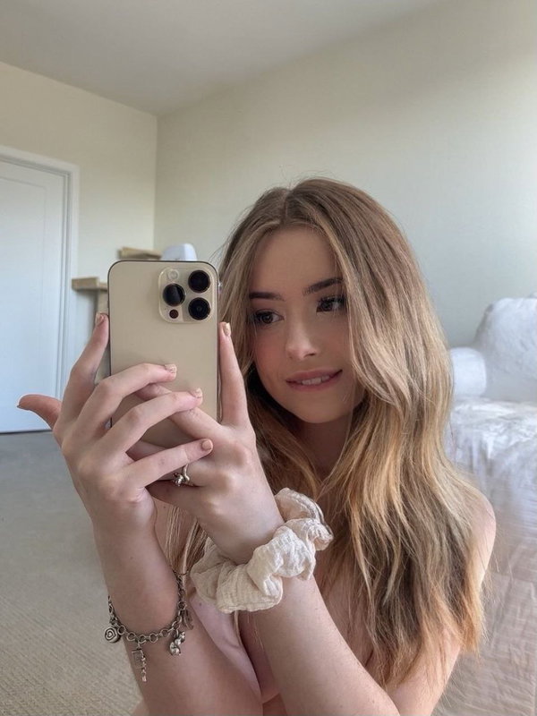 Blonde woman with long wavy hair takes a mirror selfie in a bedroom, wearing a light pink top and holding a phone.