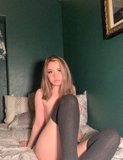 Blonde woman with shoulder-length hair sits topless on a bed, wearing dark gray leggings, against a dark green wall