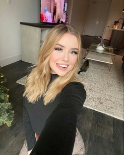 Blonde woman in a black long-sleeve top takes a selfie in a living room with a cat and Christmas tree visible.