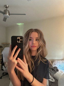 A young woman with wavy blonde hair takes a mirror selfie in a black t-shirt, standing in a bedroom with a ceiling fan