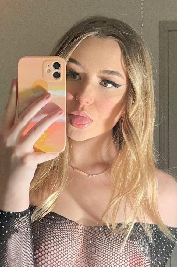 Blonde woman in a sheer black mesh top takes a mirror selfie, lips puckered, holding a peach-colored phone.
