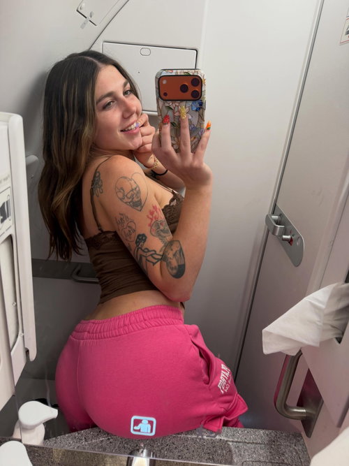 Brunette woman with multiple arm tattoos takes a mirror selfie in a public restroom, wearing a brown crop top and pink