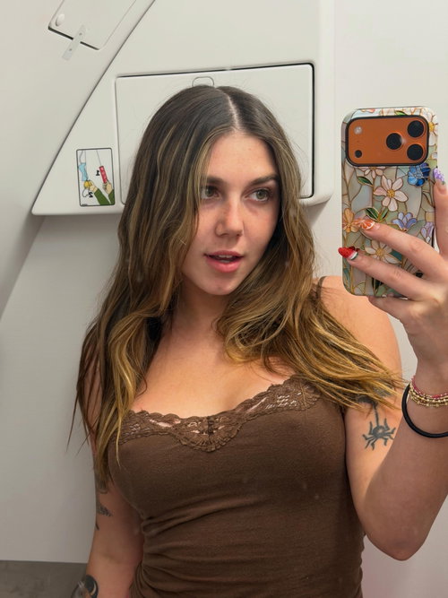 Brunette woman with wavy hair takes a mirror selfie in a brown lace-trimmed tank top, showing arm tattoos in a white