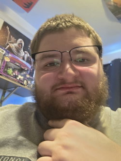 A bearded man with glasses in a gray shirt takes a close-up selfie indoors, hand near his neck, with shelves and