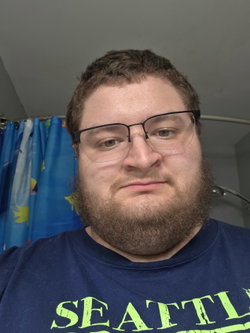 A man with a full beard and glasses wears a blue t-shirt in front of a colorful shower curtain, taking a close-up